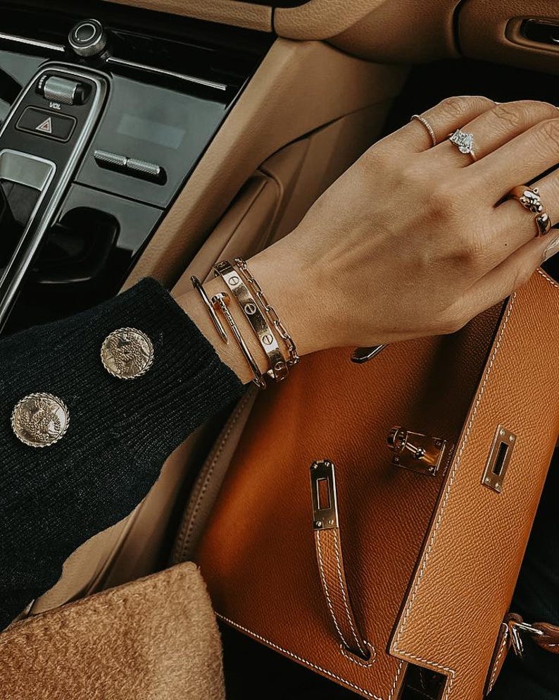 Puede incluir: Un bolso de mano de cuero marr&oacute;n claro con herrajes dorados se sostiene en un coche. La persona lleva pulseras y anillos de oro. Se ve la manga de un jersey negro con botones dorados.