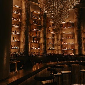 Puede incluir: Interior de un bar con poca luz y estanter&iacute;as llenas de botellas. La barra tiene una encimera curva y varias banquetas. Una l&aacute;mpara de ara&ntilde;a decorativa cuelga del techo. Las paredes son texturizadas y la iluminaci&oacute;n es c&aacute;lida.