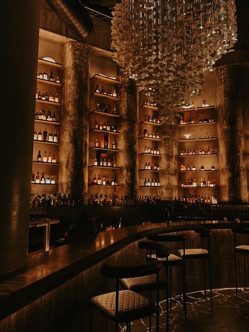 Puede incluir: Interior de un bar con poca luz y estanter&iacute;as llenas de botellas. La barra tiene una encimera curva y varias banquetas. Una l&aacute;mpara de ara&ntilde;a decorativa cuelga del techo. Las paredes son texturizadas y la iluminaci&oacute;n es c&aacute;lida.