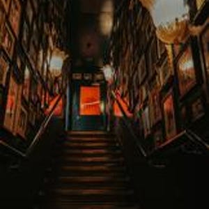 Puede incluir: Una escalera oscura asciende hacia una puerta iluminada en rojo. Las paredes est&aacute;n adornadas con numerosas fotos enmarcadas, creando un ambiente de galer&iacute;a. Dos l&aacute;mparas ornamentadas iluminan el espacio, proyectando una luz c&aacute;lida sobre los escalones de madera y las paredes oscuras.