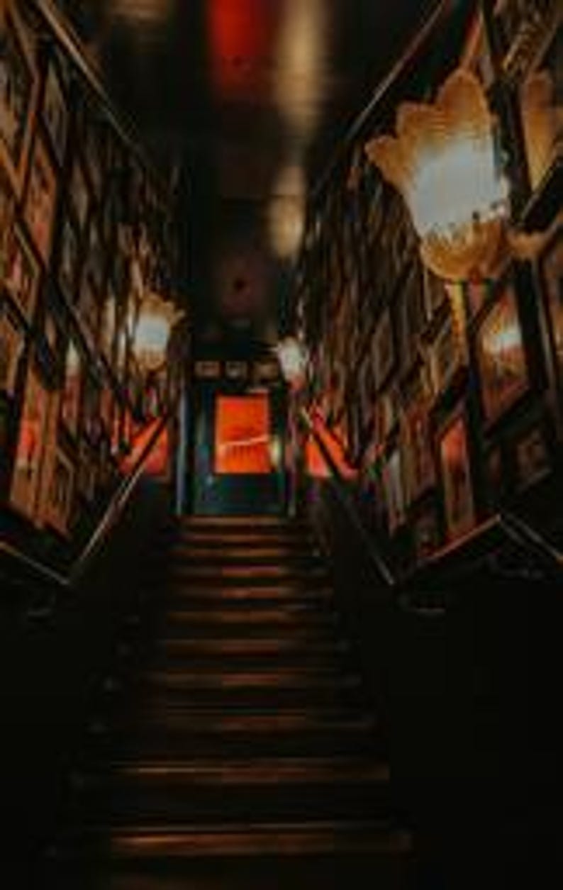 Puede incluir: Una escalera oscura asciende hacia una puerta iluminada en rojo. Las paredes est&aacute;n adornadas con numerosas fotos enmarcadas, creando un ambiente de galer&iacute;a. Dos l&aacute;mparas ornamentadas iluminan el espacio, proyectando una luz c&aacute;lida sobre los escalones de madera y las paredes oscuras.