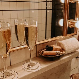 Puede incluir: Tres copas de champ&aacute;n llenas de l&iacute;quido burbujeante se encuentran sobre una superficie de m&aacute;rmol, junto a una peque&ntilde;a bandeja con productos de belleza y una toalla blanca enrollada. La escena est&aacute; ambientada en un ba&ntilde;o con un espejo dorado y un toallero de pared.