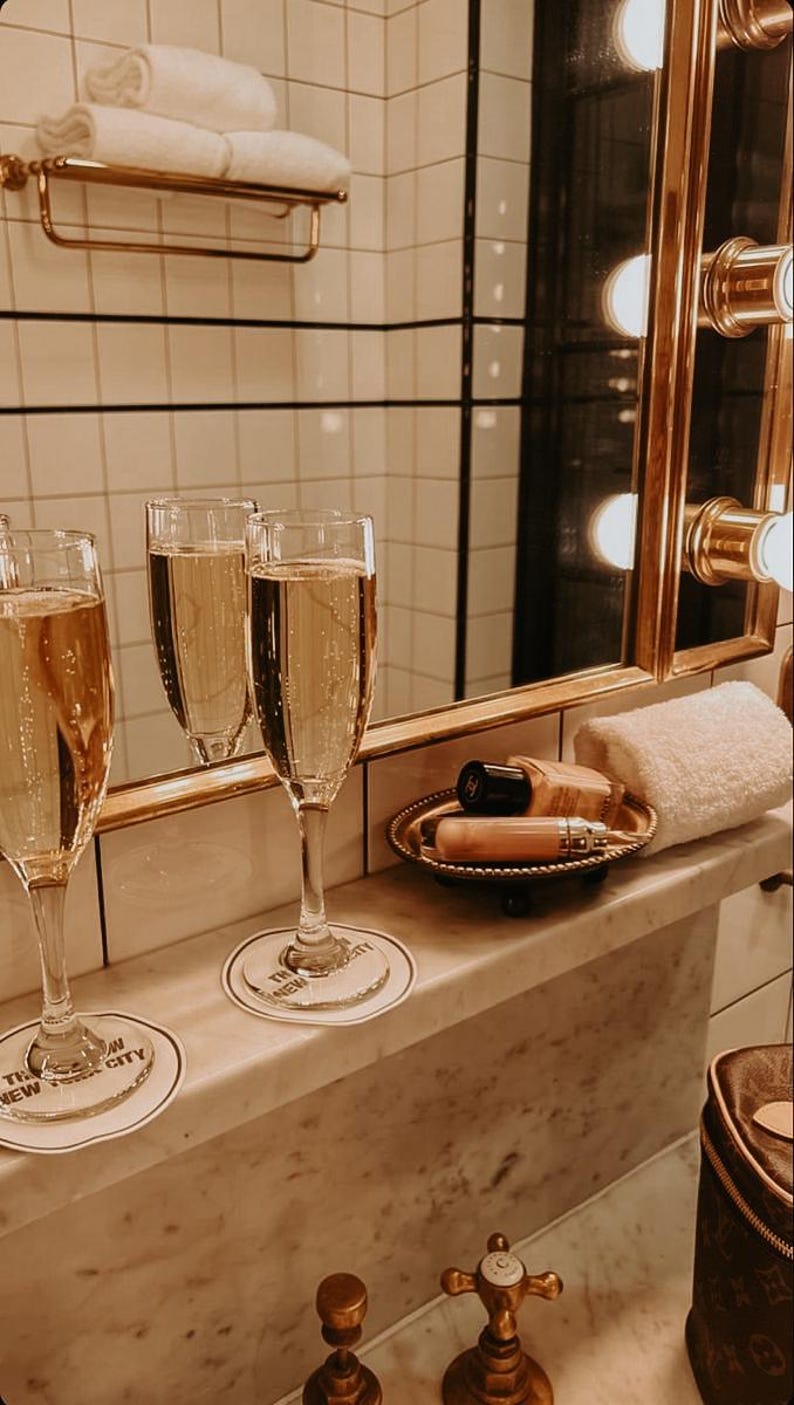 Puede incluir: Tres copas de champ&aacute;n llenas de l&iacute;quido burbujeante se encuentran sobre una superficie de m&aacute;rmol, junto a una peque&ntilde;a bandeja con productos de belleza y una toalla blanca enrollada. La escena est&aacute; ambientada en un ba&ntilde;o con un espejo dorado y un toallero de pared.