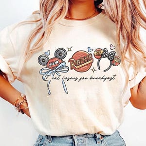 Könnte beinhalten: Ein cremefarbenes T-Shirt mit einem Grafikdruck eines roten Rennwagen-Charakters mit blauer Schleife, einem Rust-eze-Logo und Mausohren mit der Zahl 95. Der Text "I eat losers for breakfast" ist in schwarzer Schreibschrift gedruckt.