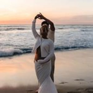 K&ouml;nnte beinhalten: Ein Paar tanzt am Strand bei Sonnenuntergang. Die Frau tr&auml;gt ein wei&szlig;es Oberteil mit langen &Auml;rmeln und einen passenden Rock. Der Mann tr&auml;gt ein helles Hemd und eine Hose. Der Ozean und der Himmel sind im Hintergrund zu sehen.