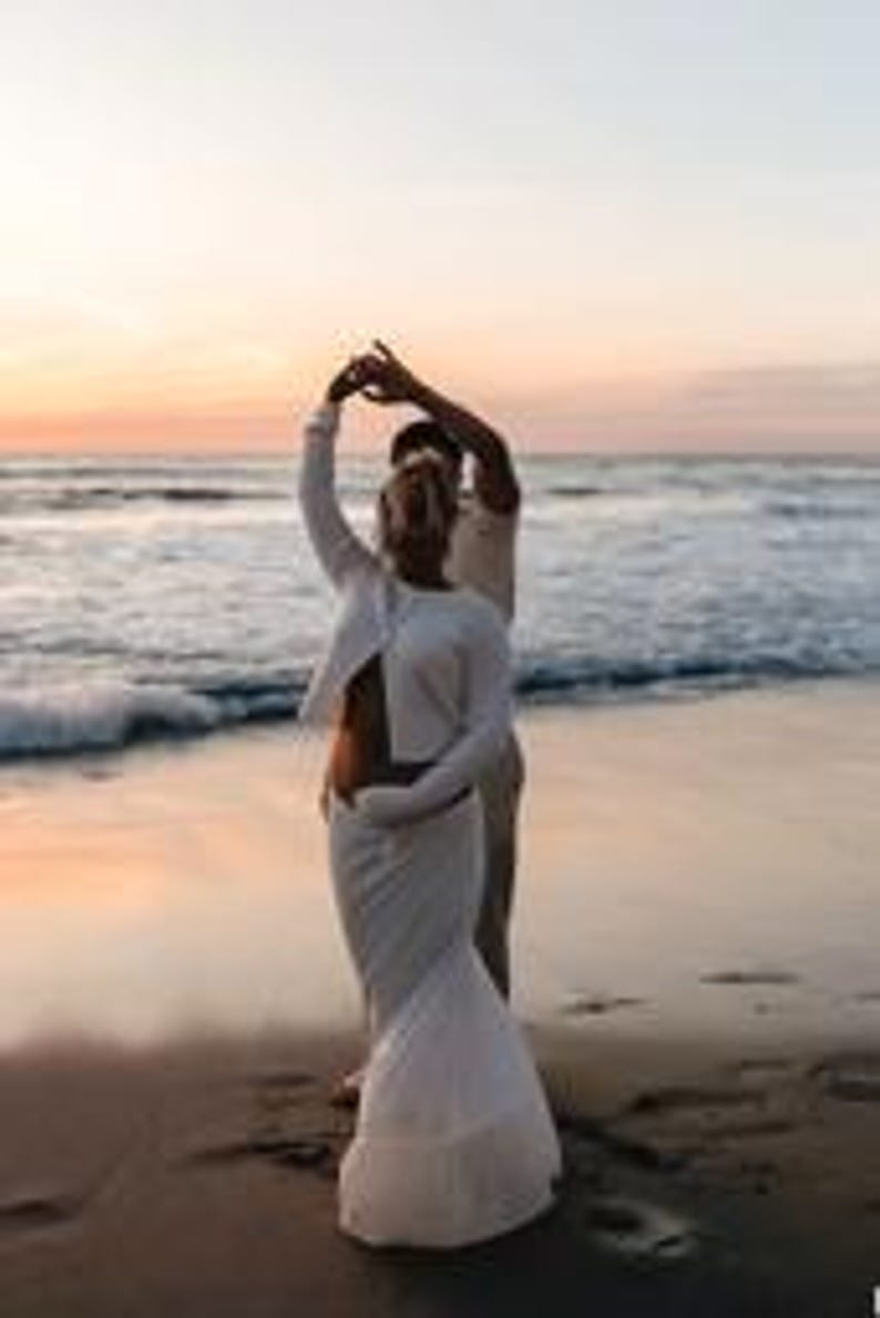 K&ouml;nnte beinhalten: Ein Paar tanzt am Strand bei Sonnenuntergang. Die Frau tr&auml;gt ein wei&szlig;es Oberteil mit langen &Auml;rmeln und einen passenden Rock. Der Mann tr&auml;gt ein helles Hemd und eine Hose. Der Ozean und der Himmel sind im Hintergrund zu sehen.