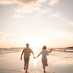 Pode incluir: Duas pessoas correm numa praia ao p&ocirc;r do sol, de m&atilde;os dadas. O c&eacute;u &eacute; laranja e amarelo quente, refletindo na areia molhada. Est&atilde;o em silhueta contra o sol poente, criando uma cena rom&acirc;ntica e alegre.