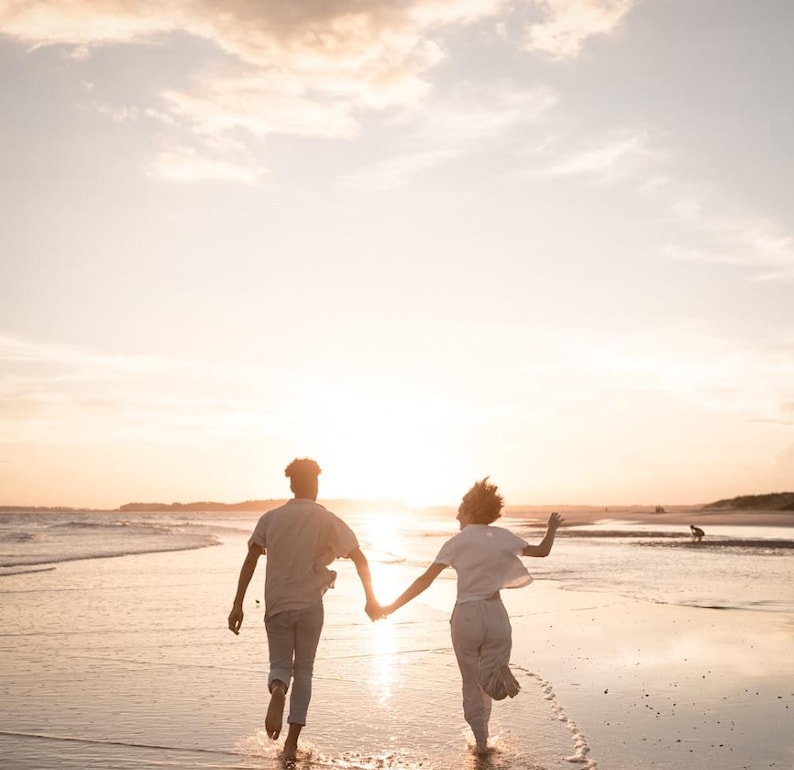 Pode incluir: Duas pessoas correm numa praia ao p&ocirc;r do sol, de m&atilde;os dadas. O c&eacute;u &eacute; laranja e amarelo quente, refletindo na areia molhada. Est&atilde;o em silhueta contra o sol poente, criando uma cena rom&acirc;ntica e alegre.