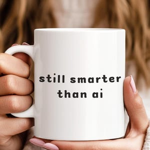 May include: A white ceramic mug featuring the black text still smarter than ai printed on the front. The mug has a standard cylindrical shape with a curved handle on the side, providing a simple and clean design for holding hot or cold beverages.