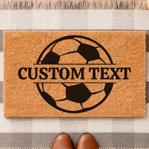 以下が含まれることがあります： 黒いサッカーボールのデザインと「CUSTOM TEXT」の文字が入った長方形のドアマット。 ドアマットは明るい茶色の素材でできており、グレーと白のチェック柄のラグの上に置かれています。 茶色のブーツが見えます。