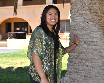 May include: A young woman with long black hair is standing in front of a palm tree. She is wearing a green and black patterned kimono-style top and has a friendly smile.