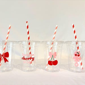 May include: Four clear plastic cups with lids and red and white striped straws. Each cup has a different cherry-themed design: a red bow, the text "She's the cherry on top", a pink bow with cherries, and a pink cherry character.