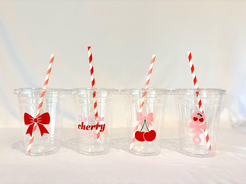 May include: Four clear plastic cups with lids and red and white striped straws. Each cup has a different cherry-themed design: a red bow, the text "She's the cherry on top", a pink bow with cherries, and a pink cherry character.