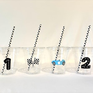 May include: Four clear plastic cups with black and white checkered straws. Each cup has a different racing-themed design: a number 1, checkered flags, a blue race car, and the number 2. Perfect for a race car themed party.