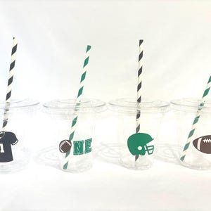 May include: Four clear plastic cups with lids, each featuring a different football-themed design. One cup has a black jersey graphic, another has the word "ONE", a green helmet, and a brown football. Each cup has a striped straw.