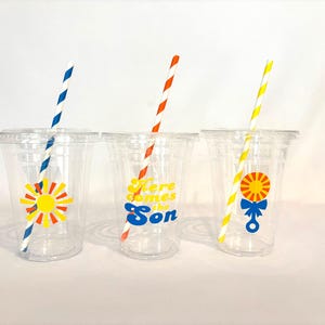 May include: Three clear plastic cups with lids and striped straws. Each cup has a different colorful graphic: a sun, the text "Here comes the Son", and a sun with a bow. The straws are blue and white, red and white, and yellow and white.
