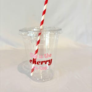 May include: A clear plastic cup with a lid and a red and white striped straw. The cup has the phrase "She is the cherry on top" printed in pink and red. The cup is designed for cold beverages.