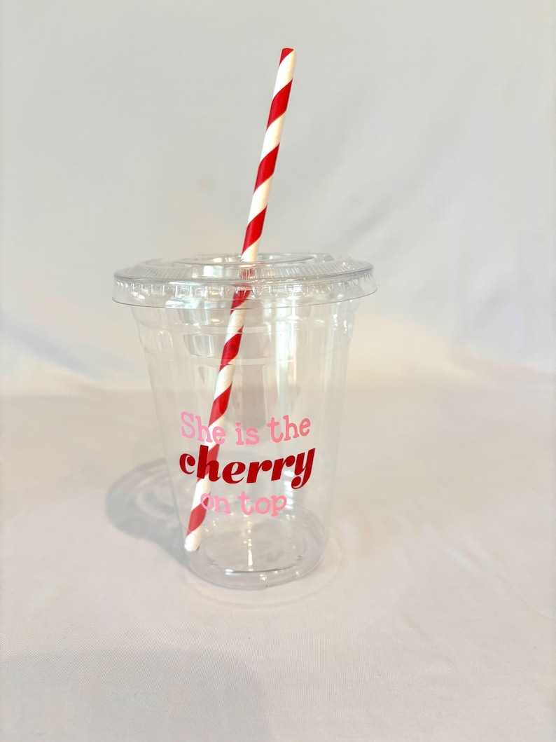 May include: A clear plastic cup with a lid and a red and white striped straw. The cup has the phrase "She is the cherry on top" printed in pink and red. The cup is designed for cold beverages.