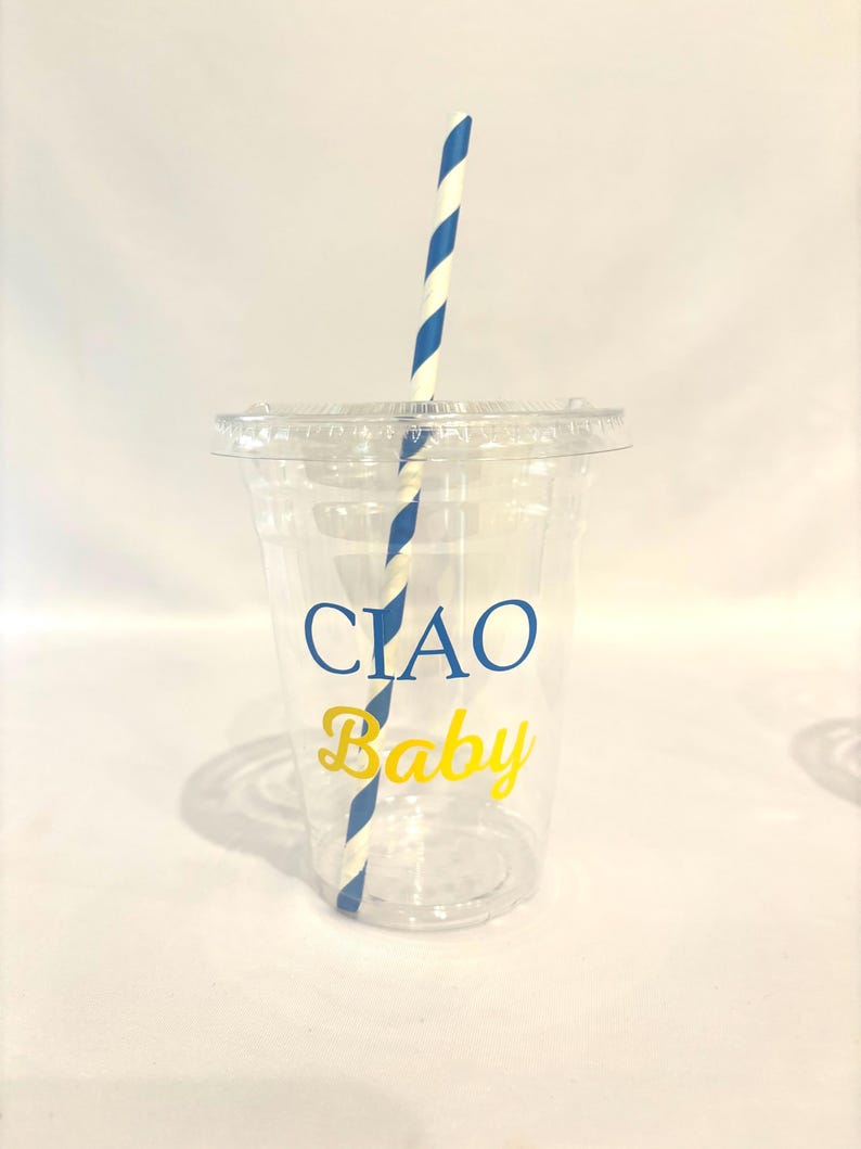 May include: A clear plastic cup with a lid and a blue and white striped straw. The cup has the words "CIAO Baby" printed on it in blue and yellow. The cup is designed for cold beverages.