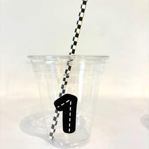 Puede incluir: Un vaso de pl&aacute;stico transparente con tapa, una pajita a cuadros blancos y negros y una pegatina negra con el n&uacute;mero uno y un dise&ntilde;o de l&iacute;nea discontinua. El vaso est&aacute; sobre una superficie blanca.