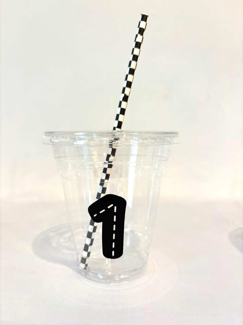 Puede incluir: Un vaso de pl&aacute;stico transparente con tapa, una pajita a cuadros blancos y negros y una pegatina negra con el n&uacute;mero uno y un dise&ntilde;o de l&iacute;nea discontinua. El vaso est&aacute; sobre una superficie blanca.