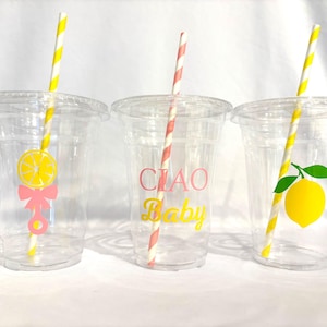 May include: Three clear plastic cups with lids and straws. Each cup has a different lemon-themed design: a lemon slice with a bow, the words "CIAO Baby", and a lemon with a leaf. The straws are striped.