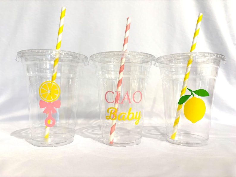 May include: Three clear plastic cups with lids and straws. Each cup has a different lemon-themed design: a lemon slice with a bow, the words "CIAO Baby", and a lemon with a leaf. The straws are striped.