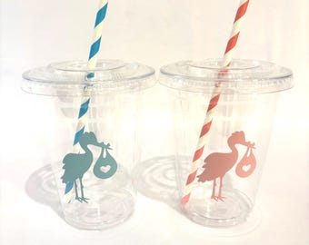 May include: Two clear plastic cups with lids and straws. Each cup has a stork graphic carrying a bundle. One stork is teal with a blue and white striped straw, the other is coral with a red and white striped straw.