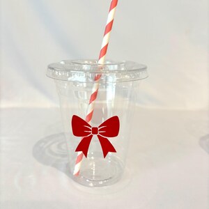May include: A clear plastic cup with a red bow design and a lid. A red and white striped straw is inserted into the cup. The cup is designed for cold beverages.