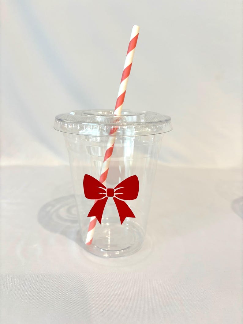 May include: A clear plastic cup with a red bow design and a lid. A red and white striped straw is inserted into the cup. The cup is designed for cold beverages.