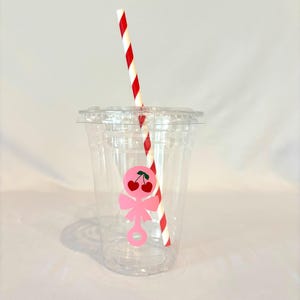 May include: A clear plastic cup with a lid and a red and white striped straw. A pink cherry design is on the cup. The cup is empty and ready to be filled with a beverage.