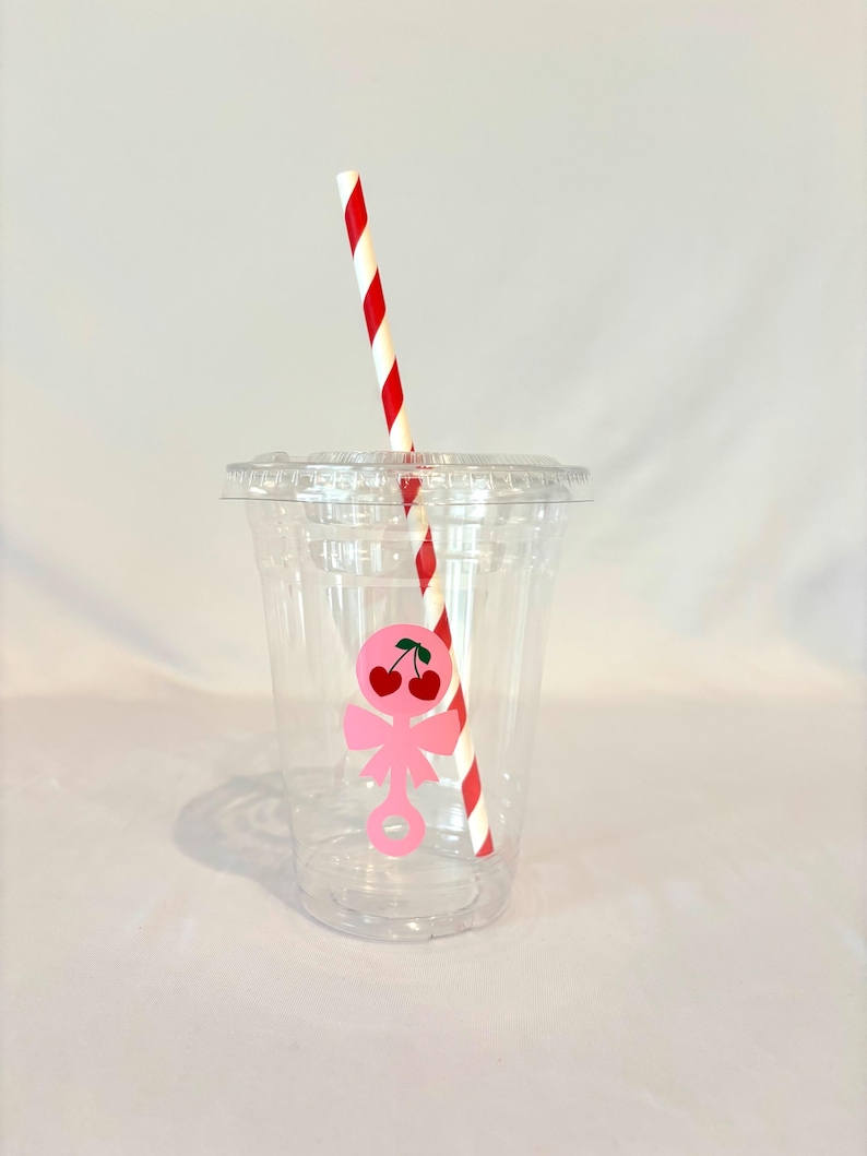 May include: A clear plastic cup with a lid and a red and white striped straw. A pink cherry design is on the cup. The cup is empty and ready to be filled with a beverage.