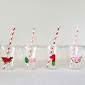 May include: Four clear plastic cups with lids, each featuring a different watermelon design. Each cup has a red and white striped straw. One cup has a watermelon slice, another has a green watermelon with the word "one", and the other two have a watermelon slice and the number "1".