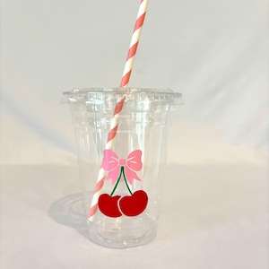 May include: A clear plastic cup with a lid and a red and white striped straw. The cup has a pink bow and two red cherries graphic. The cup is designed for cold beverages.