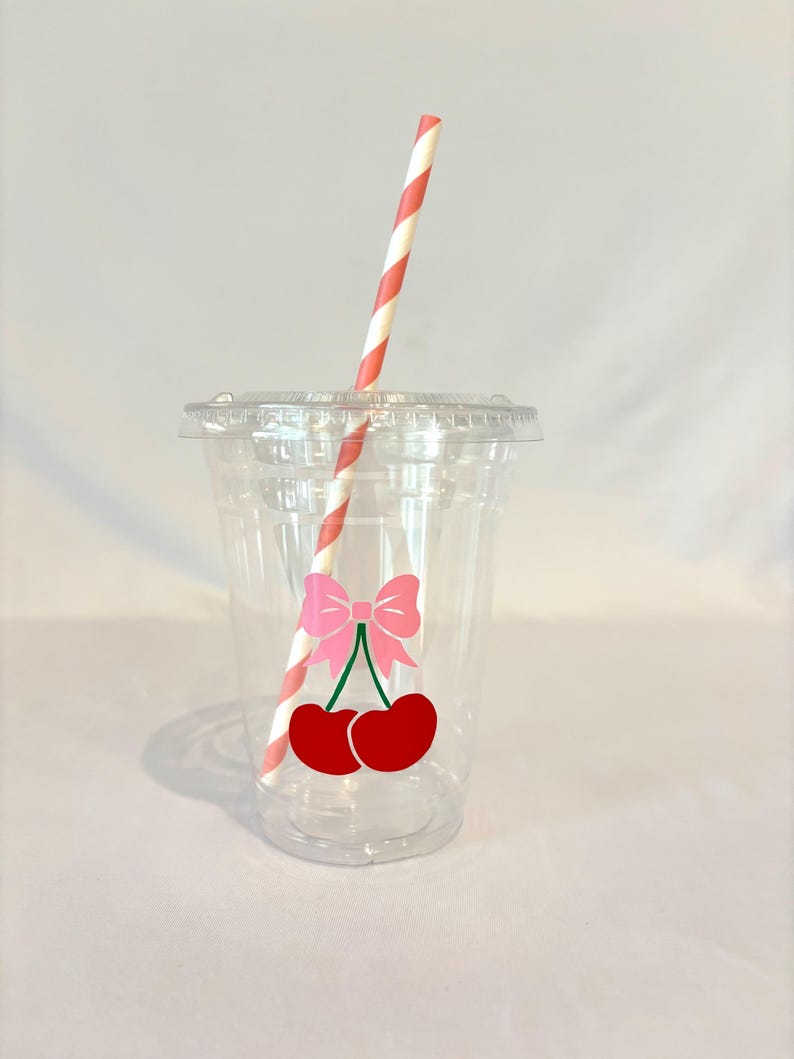 May include: A clear plastic cup with a lid and a red and white striped straw. The cup has a pink bow and two red cherries graphic. The cup is designed for cold beverages.