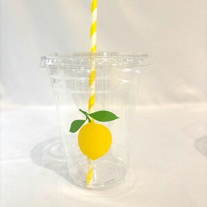 May include: A clear plastic cup with a lid, decorated with a yellow lemon graphic and green leaves. A yellow and white striped straw is inserted into the cup. The cup is designed for cold drinks.