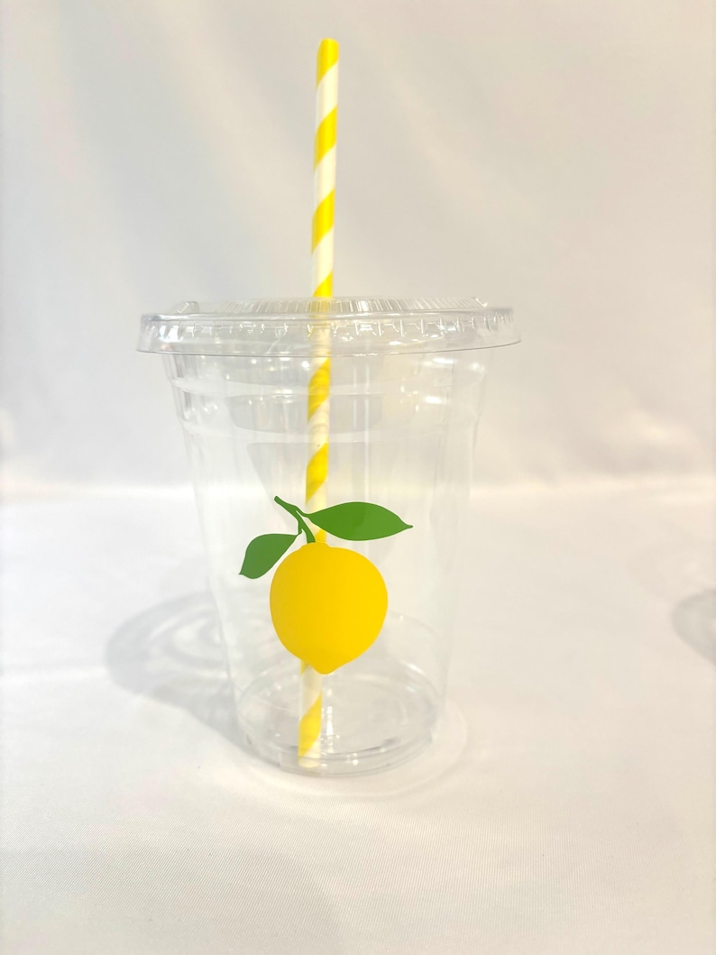 May include: A clear plastic cup with a lid, decorated with a yellow lemon graphic and green leaves. A yellow and white striped straw is inserted into the cup. The cup is designed for cold drinks.