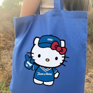 May include: Blue tote bag featuring a cartoon character wearing a baseball cap with the word "LOGO" and a red bow. The character is holding up a foam finger with the number "1" and the words "Team Name" on the jersey. The bag is held by a person.