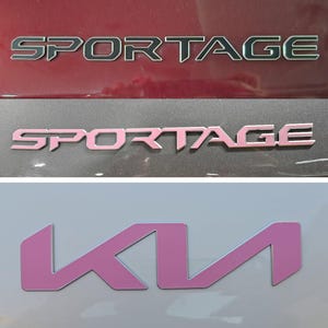 May include: A collection of vehicle emblems featuring the word SPORTAGE in a metallic finish on a dark red surface, a light pink SPORTAGE emblem on a dark gray surface, and a light purple KIA logo on a light gray surface.