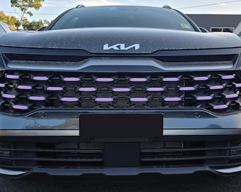 May include: The front view of a dark gray Kia vehicle featuring a prominent silver Kia logo on the hood. The black grille is accented with horizontal light purple rectangular inserts arranged in a repeating pattern across the front fascia.