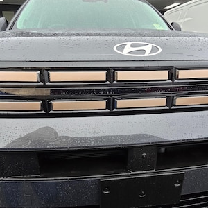 May include: Close-up of a dark gray Hyundai vehicle's front end. The grille features horizontal bronze-colored accents. The Hyundai logo is centered above the grille. The bumper is black, and the vehicle appears to be wet.