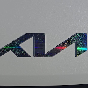 May include: A close-up of a car emblem. The emblem features the letters "K" and "I" in a stylized design. The letters are black with a holographic, rainbow-coloured effect. The emblem is on a light grey surface.