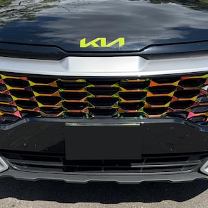 May include: Close-up of a black car's front grille with a vibrant, multi-coloured honeycomb pattern. The Kia logo in neon yellow is visible above the grille. The car's bumper and headlights are also visible.