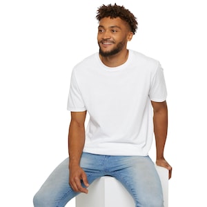 May include: A white t-shirt with a crew neck and short sleeves. The person is wearing blue jeans and sitting on a white surface.