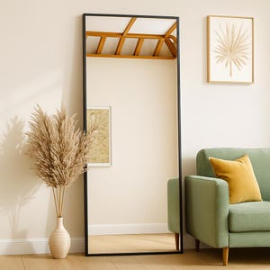 May include: A full-length rectangular mirror with a black frame leans against a wall. A vase of dried pampas grass sits in front of the mirror. A green sofa with a yellow pillow is to the right. A framed picture hangs on the wall.