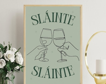 May include: A framed print with a sage green background features the word "SLÁINTE" above and below a line drawing of two wine glasses clinking. The glasses are held by hands, suggesting a toast. The frame is a light wood color.