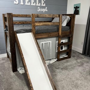Könnte beinhalten: Ein hölzernes Etagenbett mit eingebauter Rutsche. Der Bettrahmen ist dunkelbraun gebeizt, mit einer weißen Rutsche. Das obere Bett hat graue Bettwäsche. Die Namen "STEELE" und "Joseph" sind an der Wand über dem Bett zu sehen.