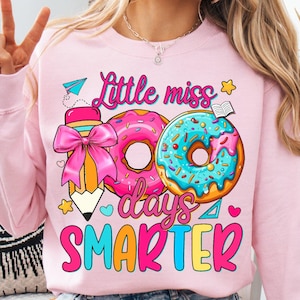 May include: Pink sweatshirt with a colorful graphic design. The design features the text "Little miss 100 days Smarter" with illustrations of donuts, a pencil, and a book. The donuts are pink and blue with sprinkles.