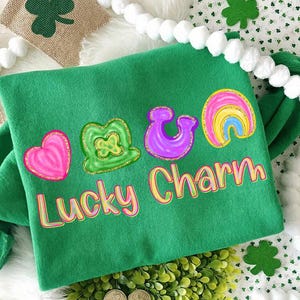 May include: A green sweatshirt with a "Lucky Charm" design featuring a pink heart, a green shamrock, a purple horseshoe, and a rainbow. The design has a yellow and pink outline. The sweatshirt is surrounded by decorative elements.