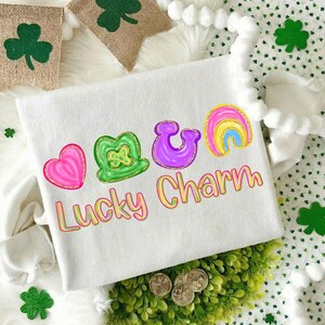 May include: White sweatshirt with a colorful "Lucky Charm" design featuring a pink heart, green shamrock, purple horseshoe, and rainbow. The design has a glittery outline. Decorative shamrock banners and accents are in the background.